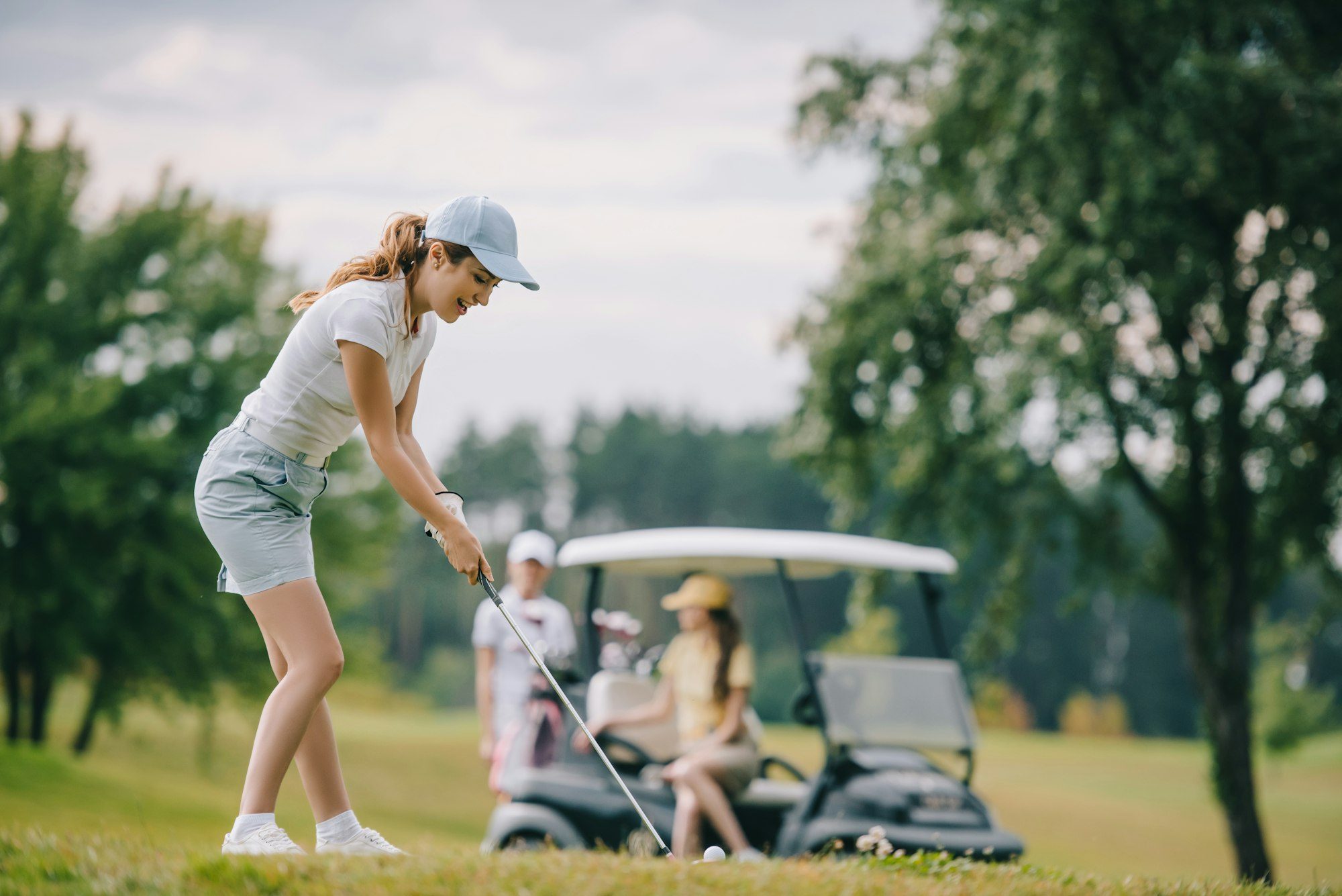 selective focus of woman with golf club playing golf and friends resting at golf cart on green lawn